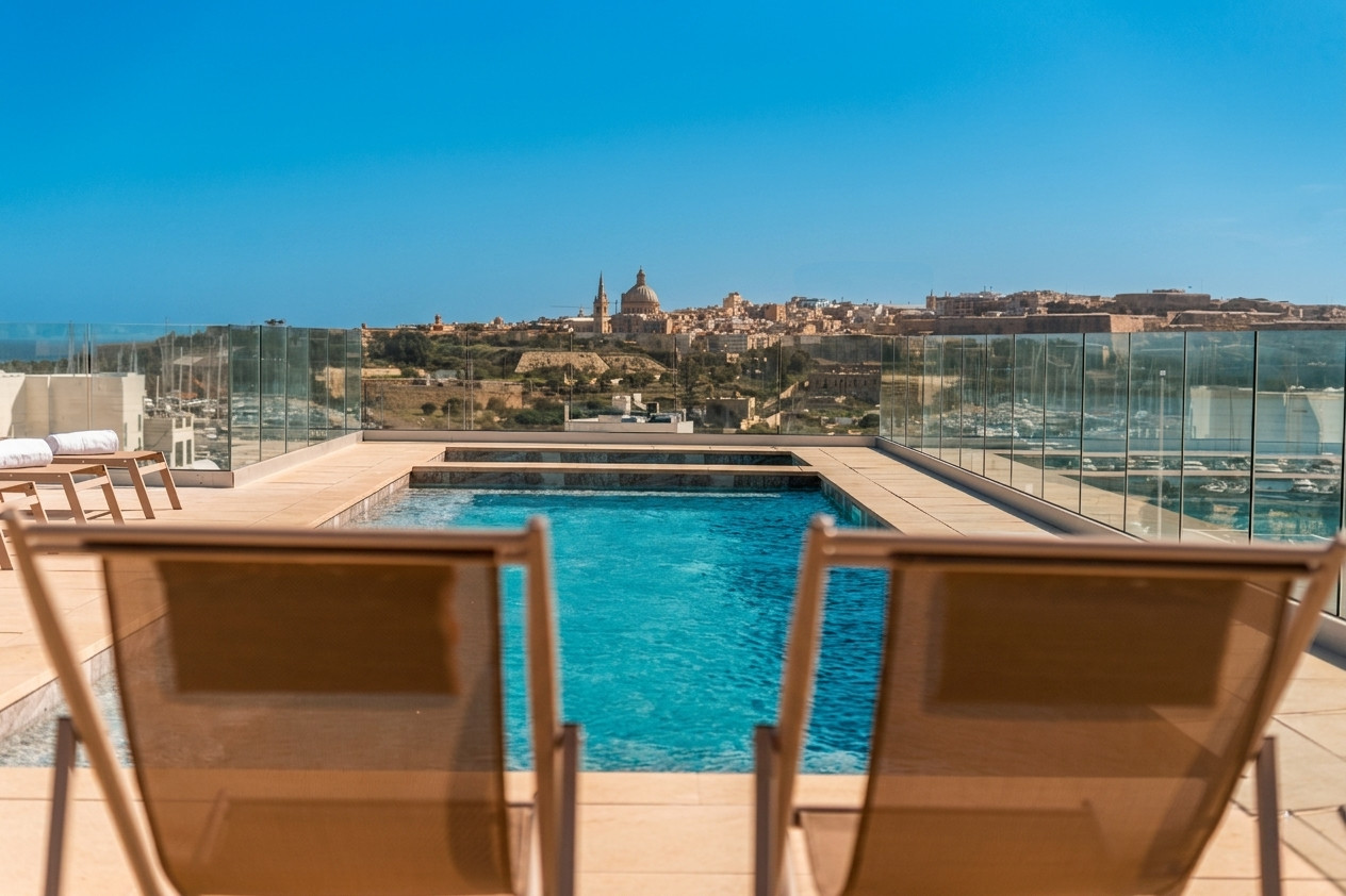 Rooftop pool with sun loungers overlooking Valletta’s skyline, including the domed St. Paul’s Cathedral, under a clear blue sky.